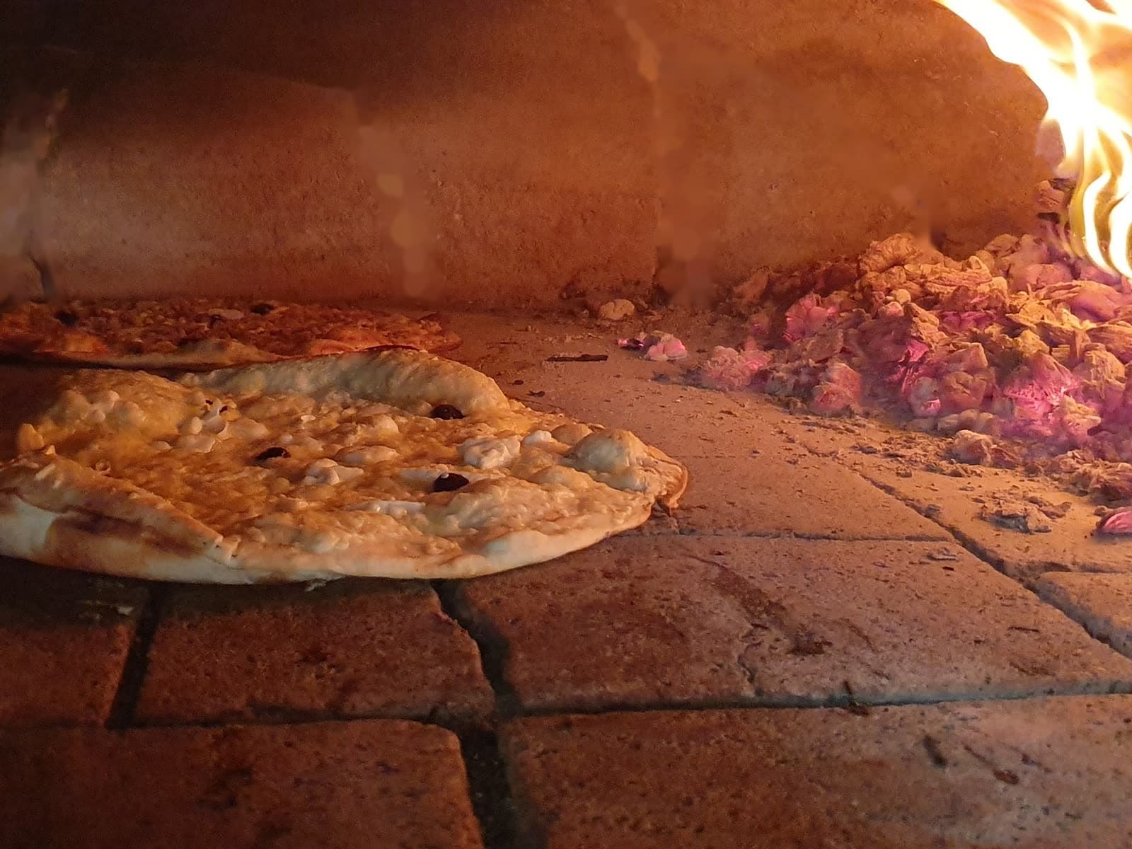 Deux pizzas en train de cuire dans le four à bois, braises et flammes au fond